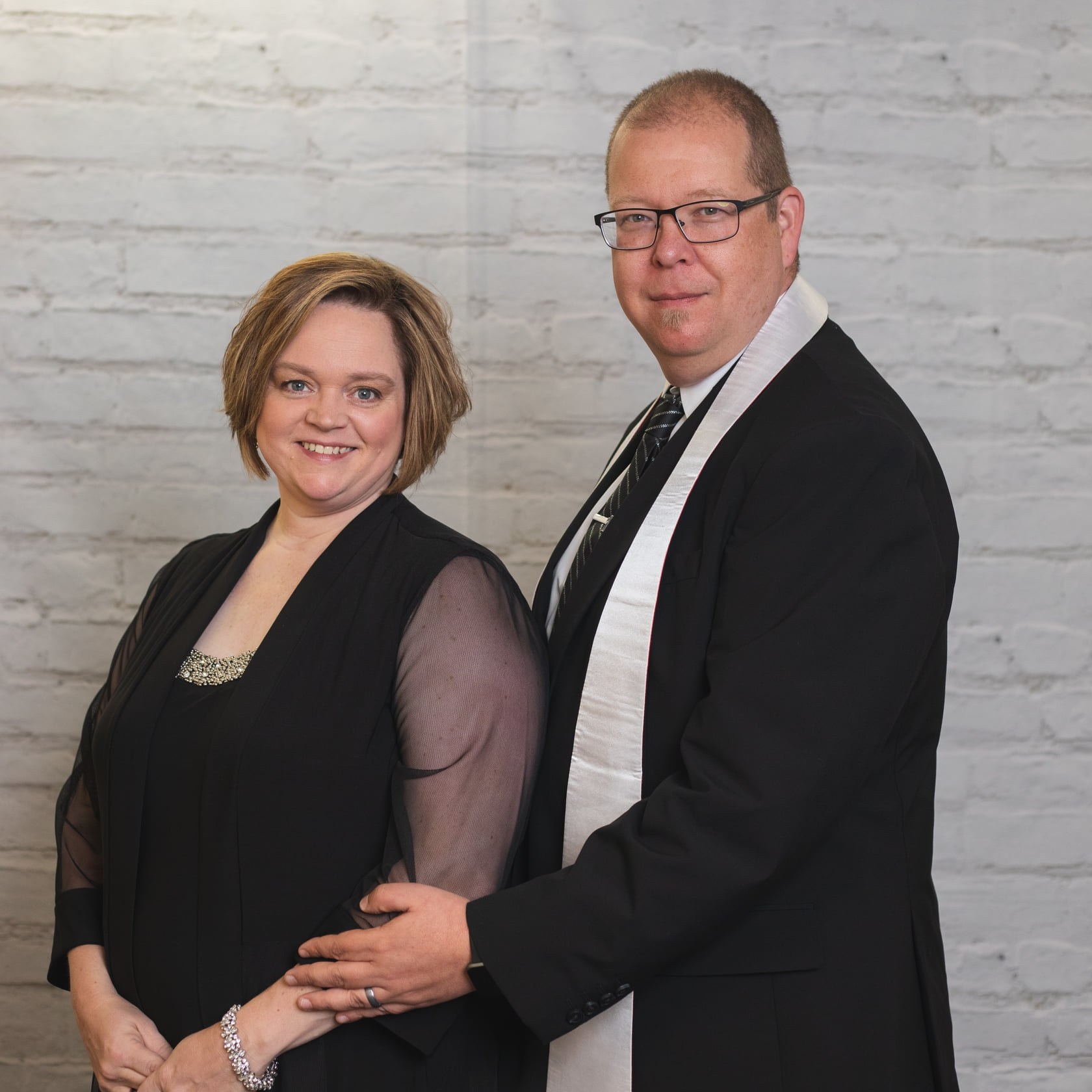 A smiling couple standing together in front of a light brick wall, dressed in formal black attire, facing the camera with their arms gently linked. The woman has short hair and a sheer-sleeved black top with a decorative neckline, and the man is wearing glasses, a suit, and a light-colored stole over his shoulder.